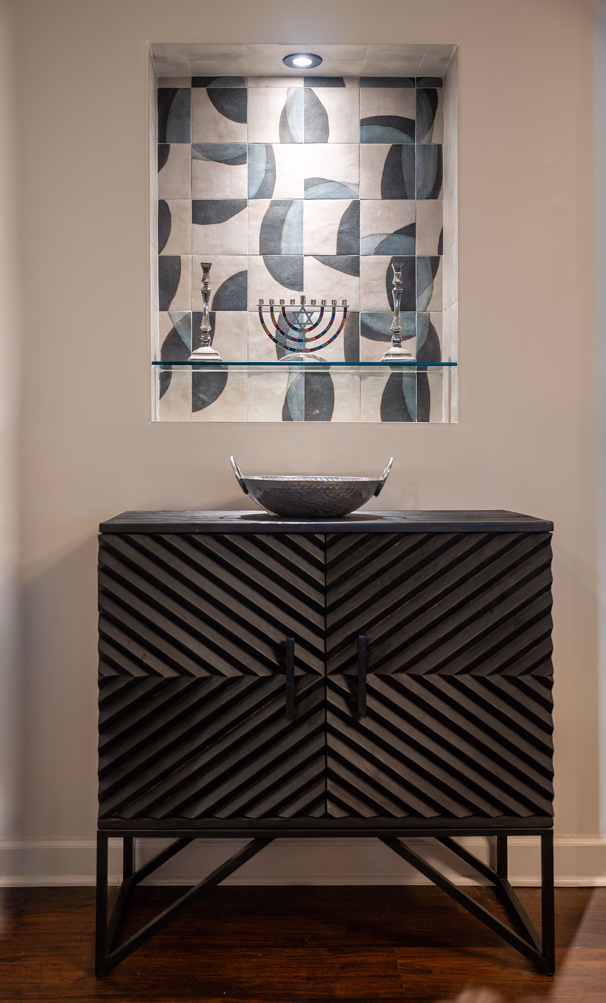 Entryway console with a dark chevron-front cabinet, vessel sink bowl, and a lit display niche with Judaica pieces