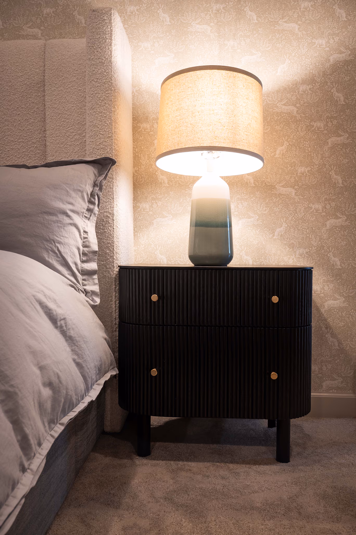 Primary bedroom nightstand with a ceramic table lamp and subtle rabbit-print wallpaper