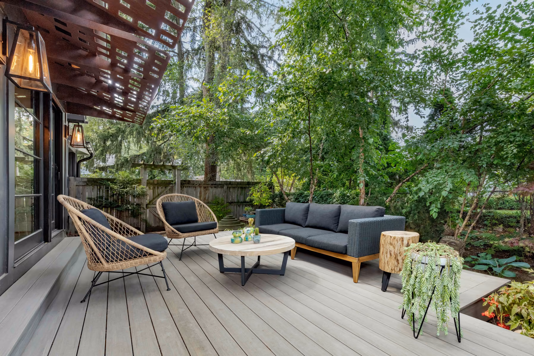 Covered deck with a wood pergola, wicker outdoor chairs, and a gray sectional sofa surrounded by greenery