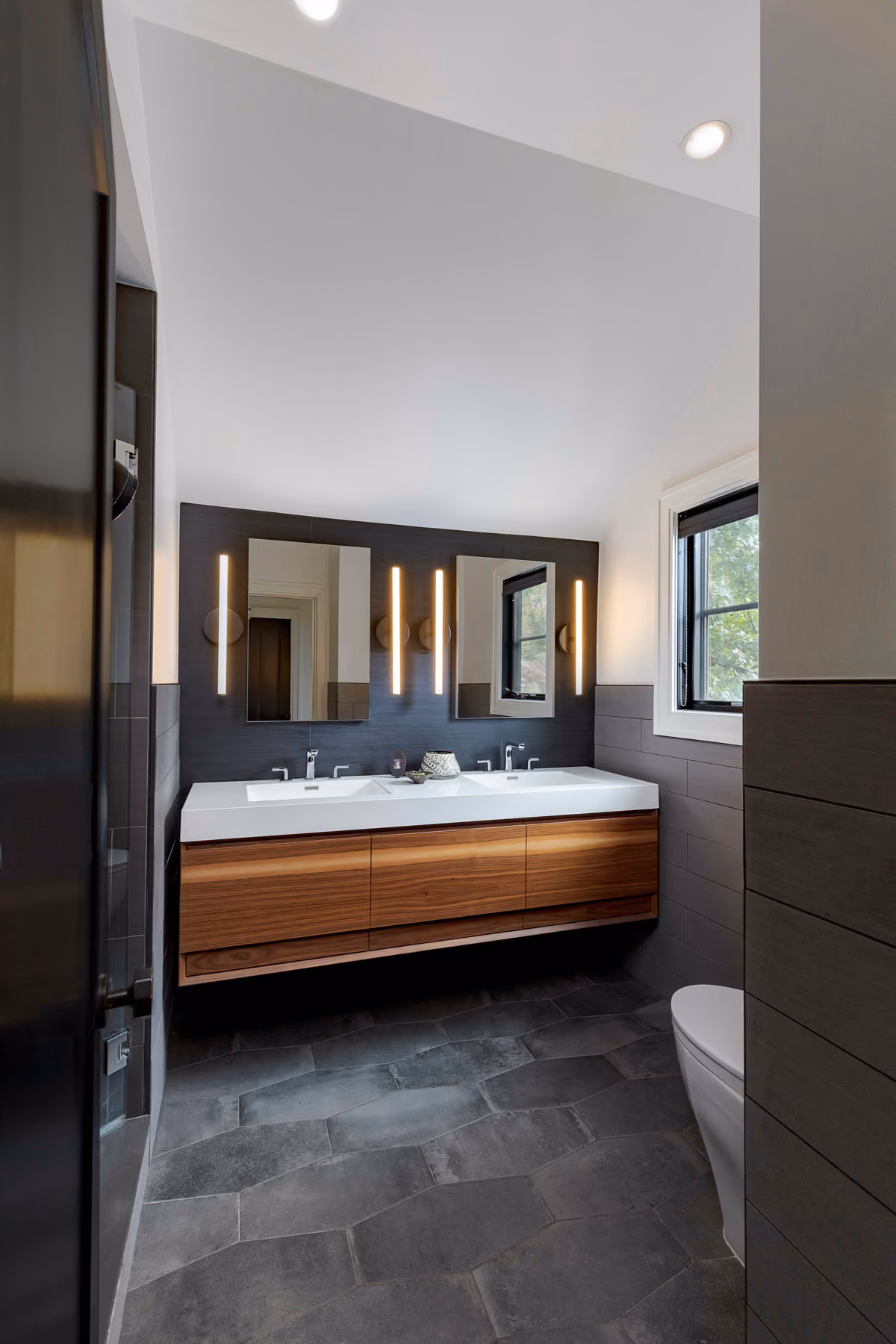 Bathroom with a floating walnut double vanity, dark stone tile, and vertical brass tube sconces