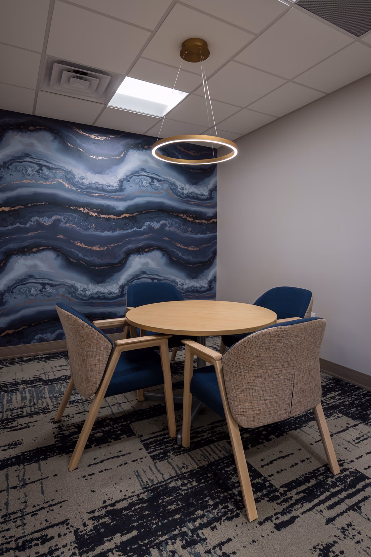 Consultation room with a wave-patterned accent wall, round wood table, and mixed navy and gray chairs under a circular pendant light