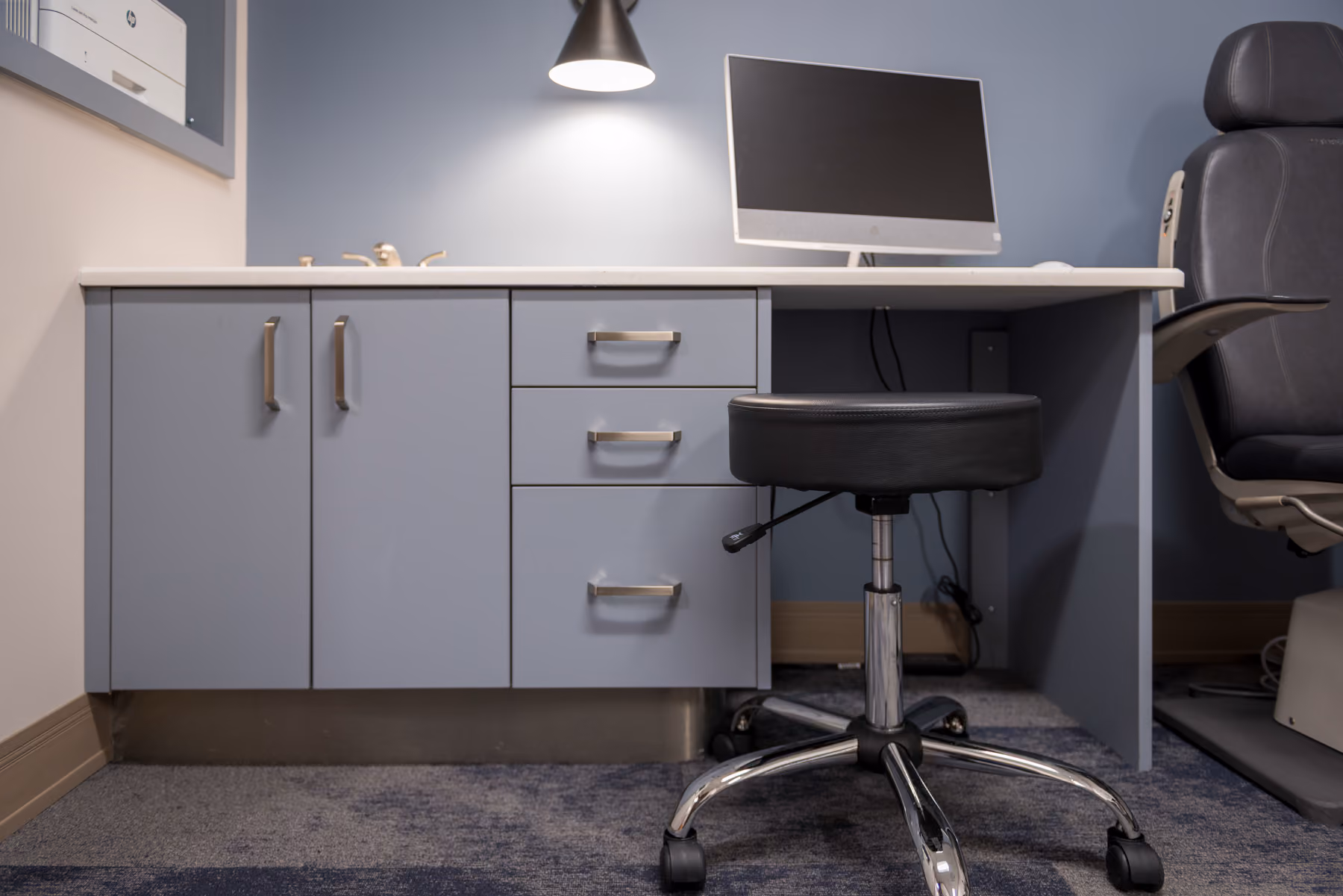 Exam room with light blue-gray cabinetry, a built-in computer desk, and a black exam chair