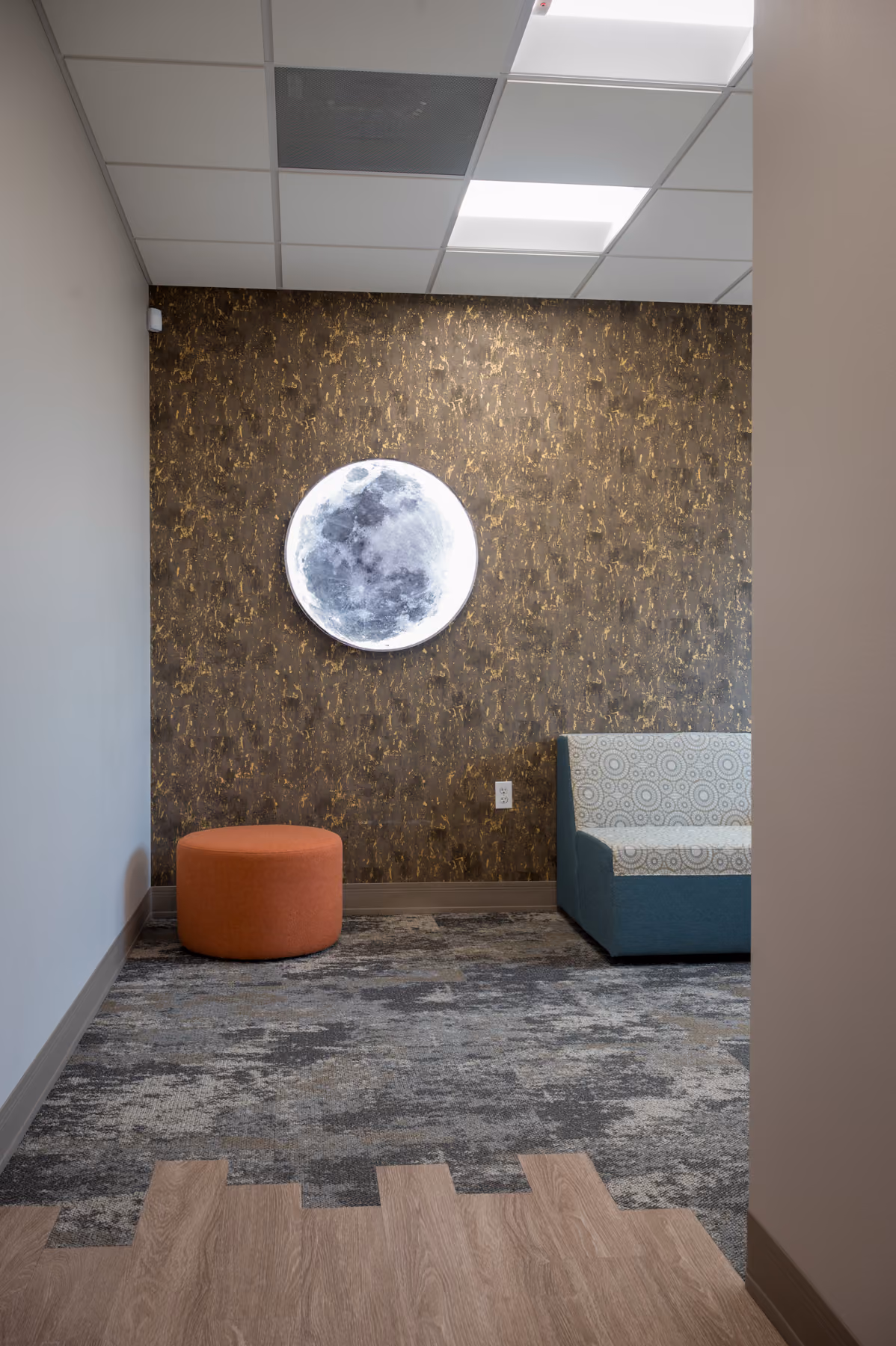 Wide view of the waiting nook with moon wall art and a mix of wood-look flooring and patterned carpet tile
