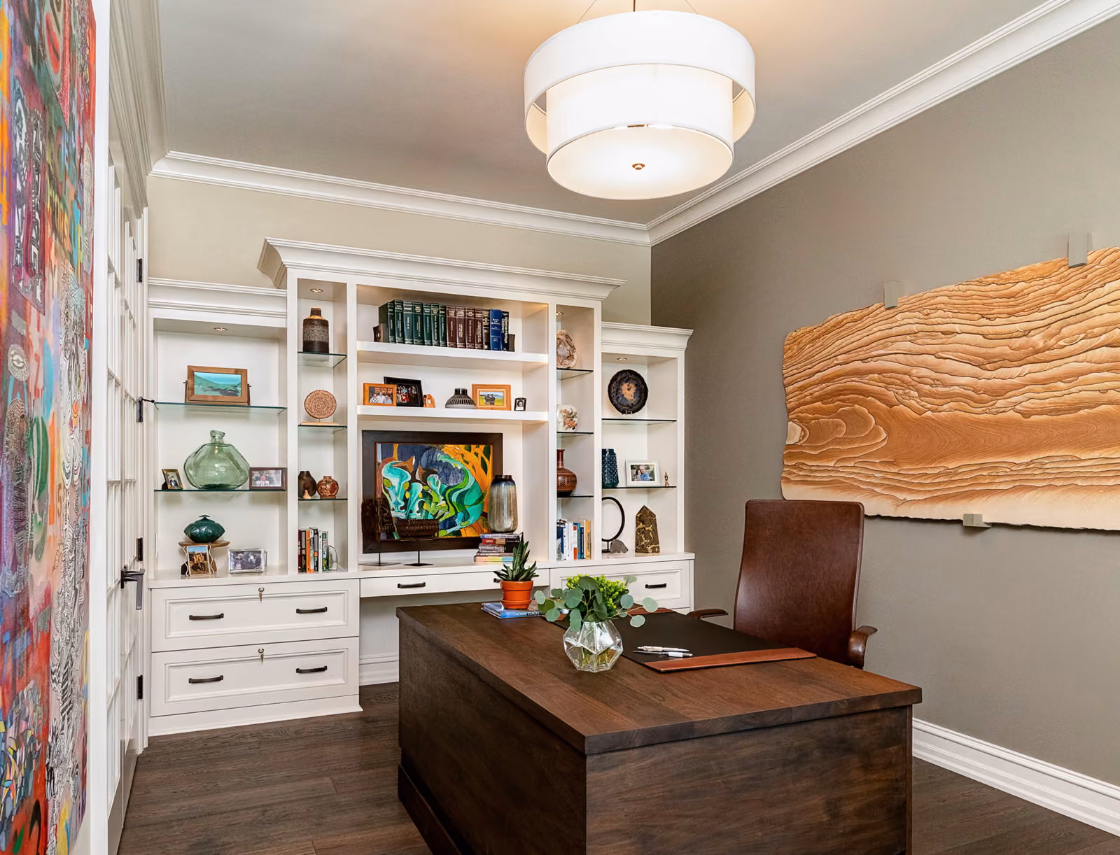 Home office with white built-in bookshelves displaying curated art-glass objects, a dark wood desk, and live-edge wood wall art