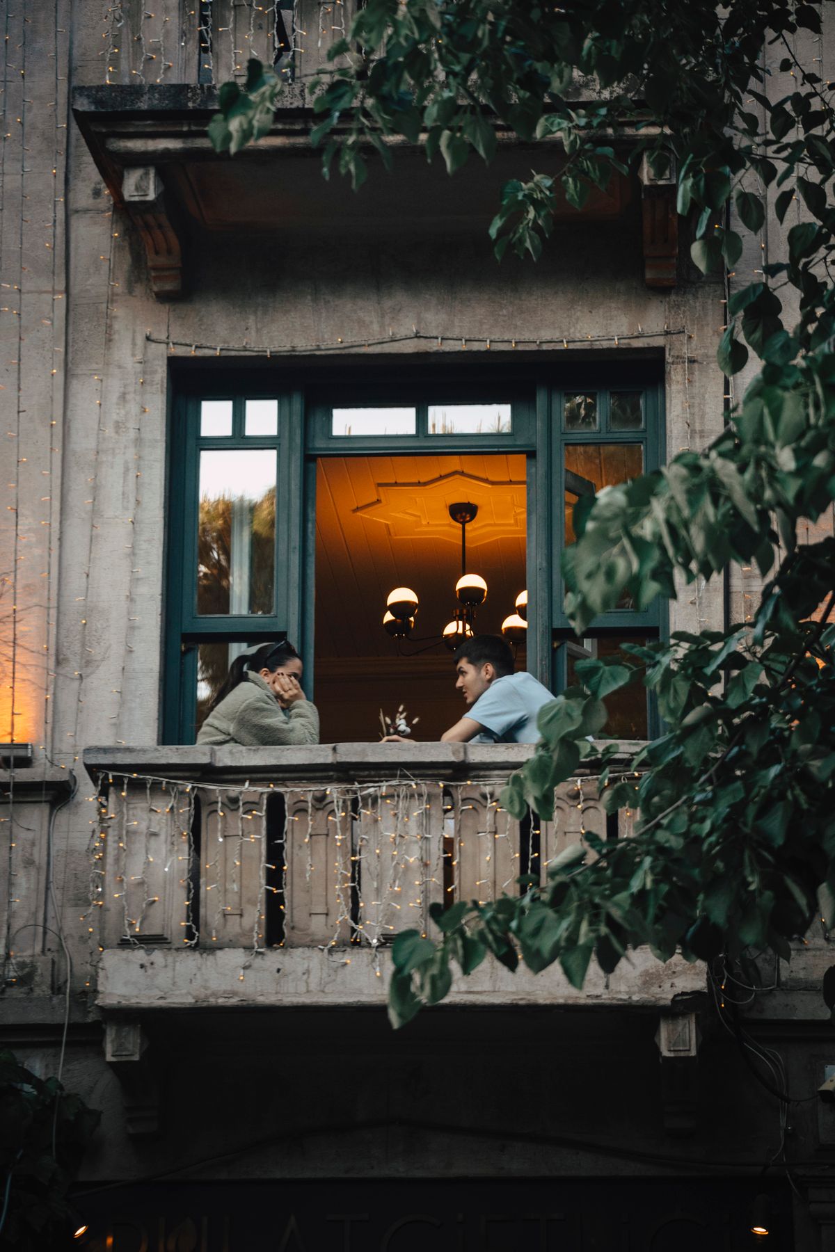 Two people talking on a balcony strung with fairy lights