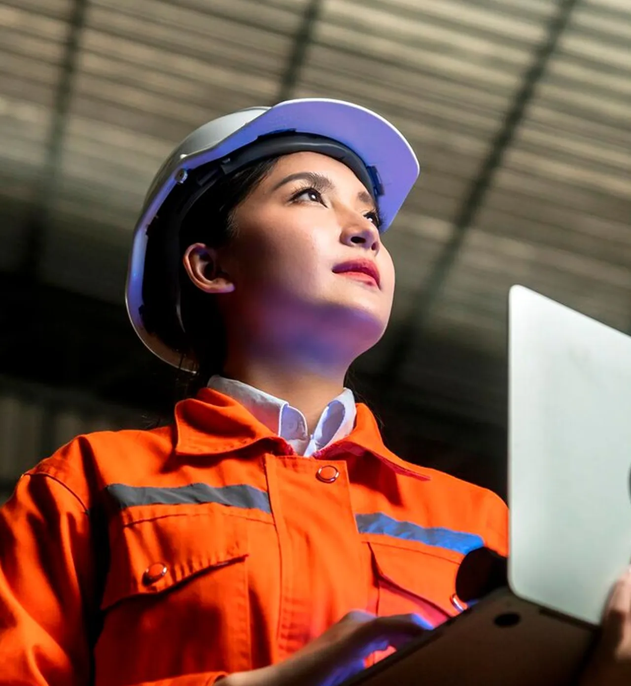 Woman engineer in an orange safety jacket and white hard hat working on a laptop indoors.