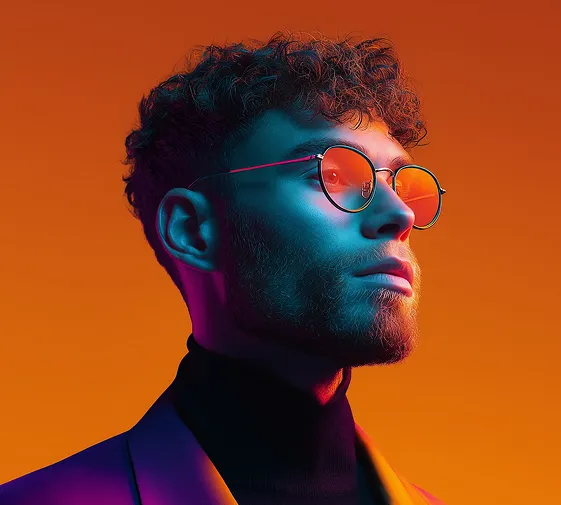 Side profile of a young man with curly hair wearing round glasses and a dark turtleneck against orange.