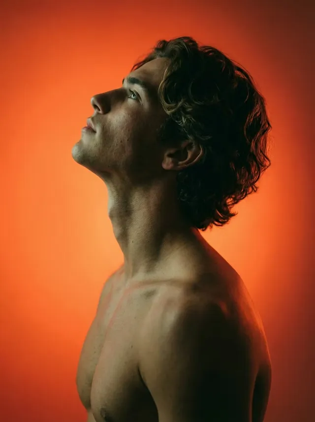 Side profile of shirtless young man with curly hair looking upward against an orange background.