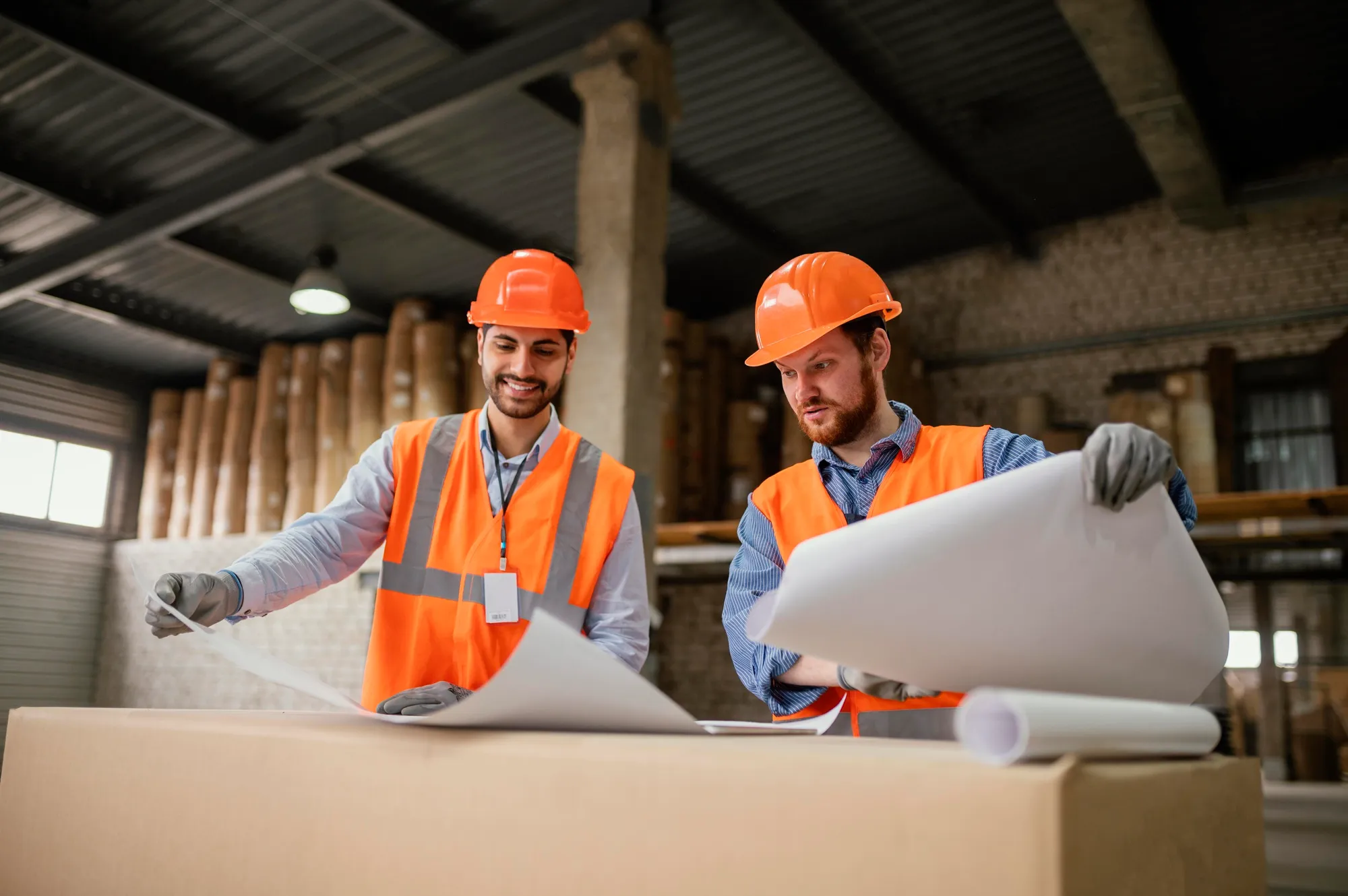 Two construction workers in orange vests and helmets reviewing blueprints in a warehouse.