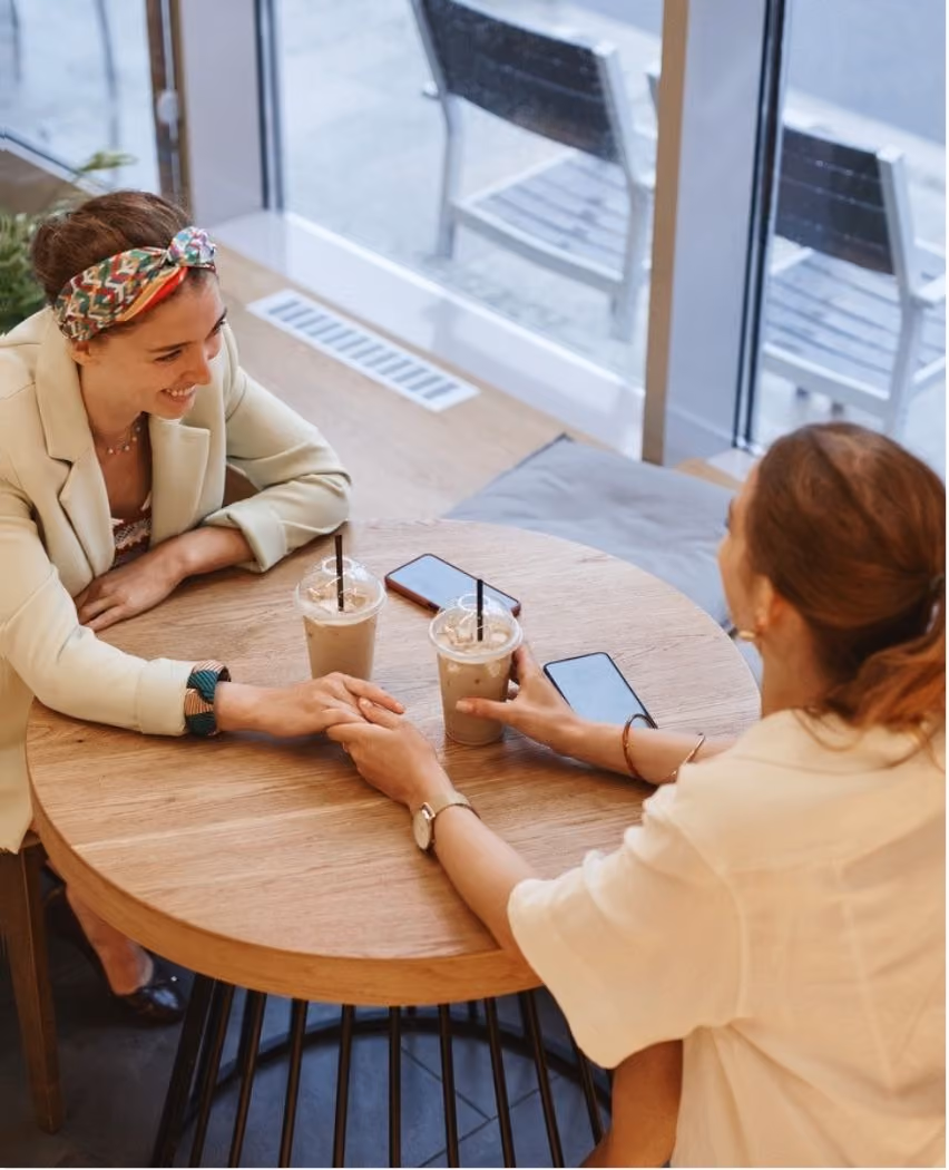 Two women sitting at a round wooden table indoors, holding hands and smiling, each with an iced coffee and a smartphone on the table.