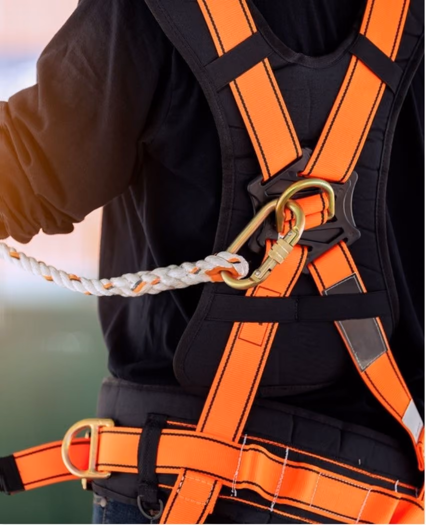 Close-up of a person wearing an orange safety harness with straps crossing on the back, attached to a white and orange braided safety rope via a gold carabiner clip.