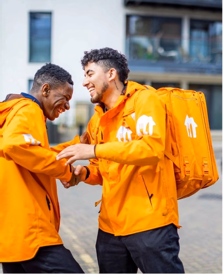 Two delivery workers in bright orange jackets and backpacks shake hands and laugh outdoors in an urban setting.