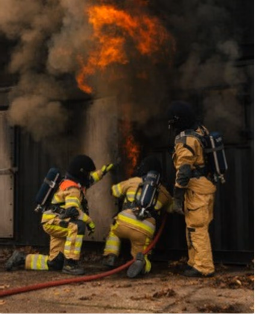 Three firefighters in full gear, including breathing apparatus, actively working to extinguish flames and thick black smoke coming from a dark structure, with a red hose on the ground.