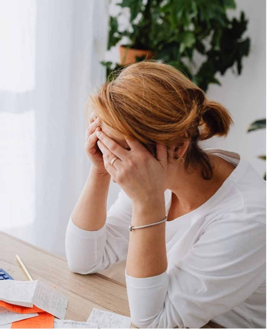 Woman sitting at a desk with her hands covering her face, appearing stressed or overwhelmed, with scattered papers and a pen in front of her and a green plant in the background.