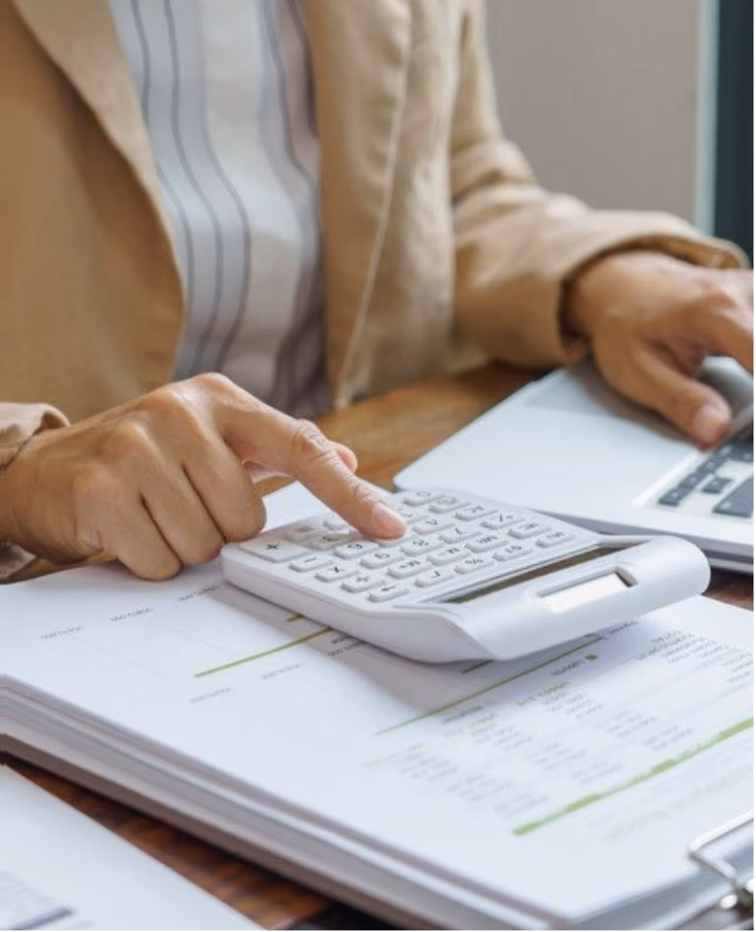 Person in a beige jacket using a white calculator and a laptop on a wooden desk with printed documents underneath.