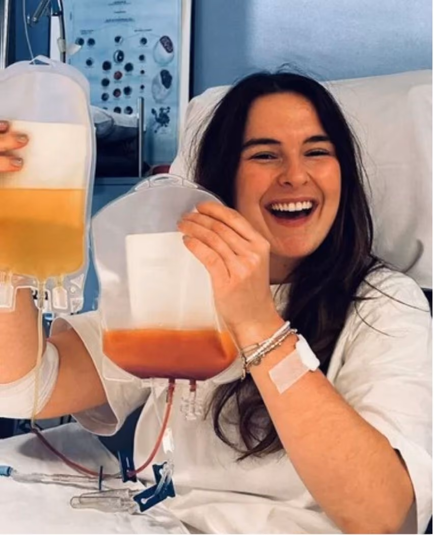A woman in a hospital bed smiling and holding up two bags of blood plasma, one yellow and one red, connected to her arm via IV lines.
