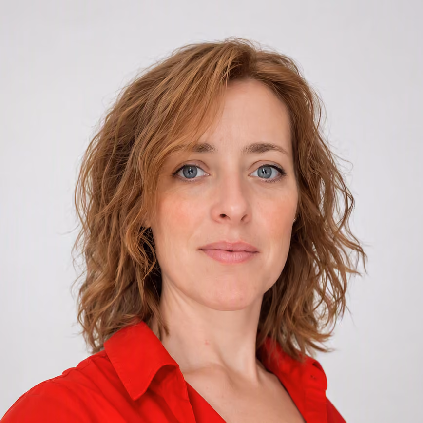 Close-up portrait of a woman with wavy light brown hair and blue eyes, wearing a red collared shirt, looking confidently at the camera against a plain light background.