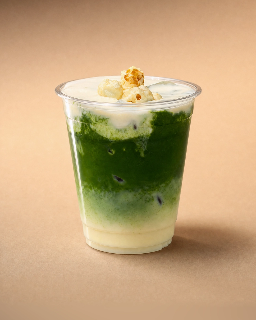 Corn matcha, pale corn milk at the base and a deep band of green matcha above it, under a white popcorn cold foam crowned with caramel popcorn.