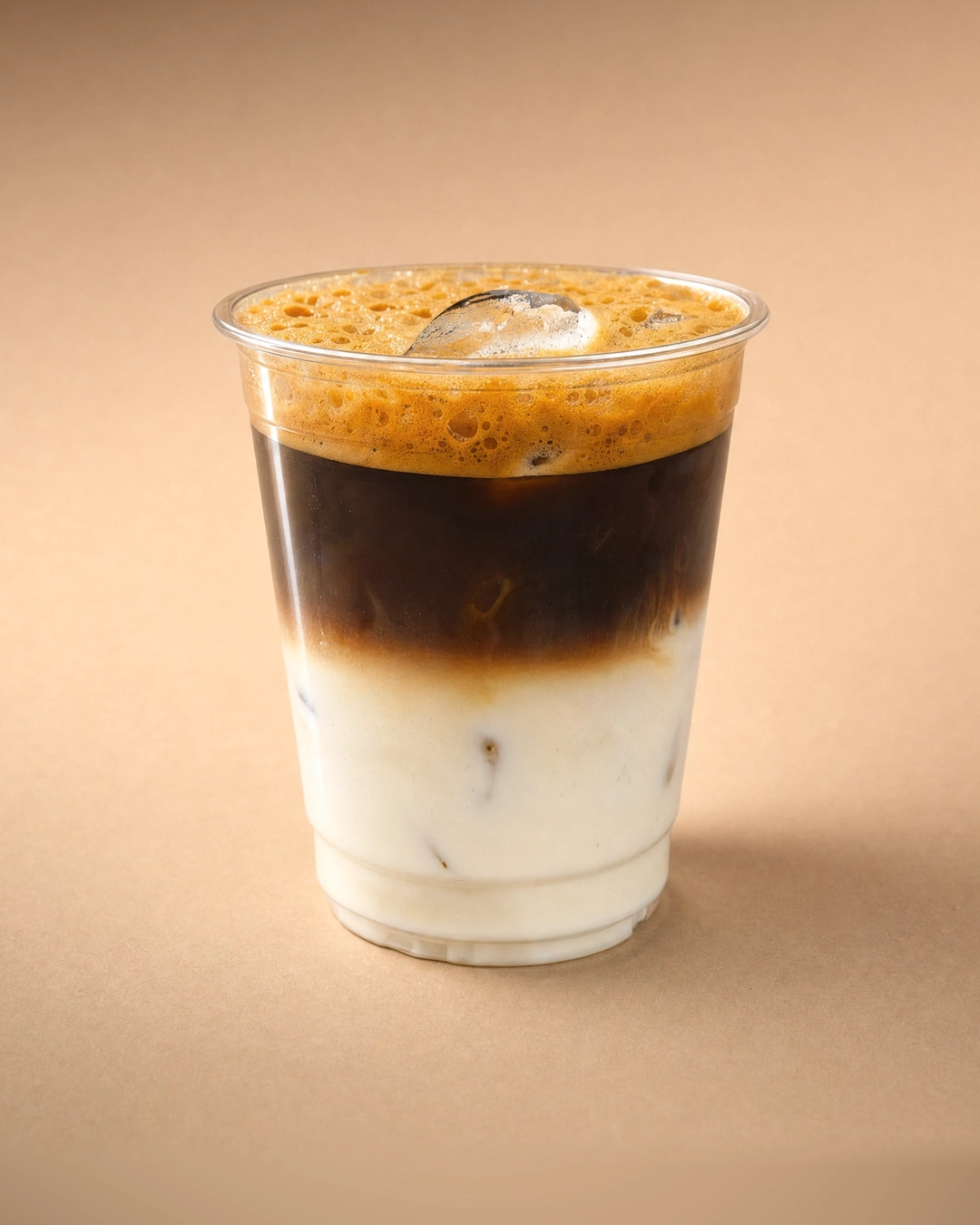 Bạc xỉu (iced latte) in three bands: white milk and ice at the bottom, near-black coffee above, and a coarse orange-tan crema with an ice cube poking through.