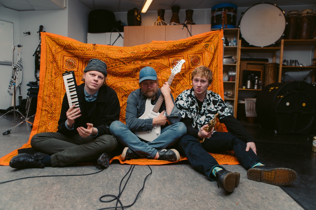 Three guys sitting in a studio. One with a small keyboard, one with a guitar and one with a saxophone. 
