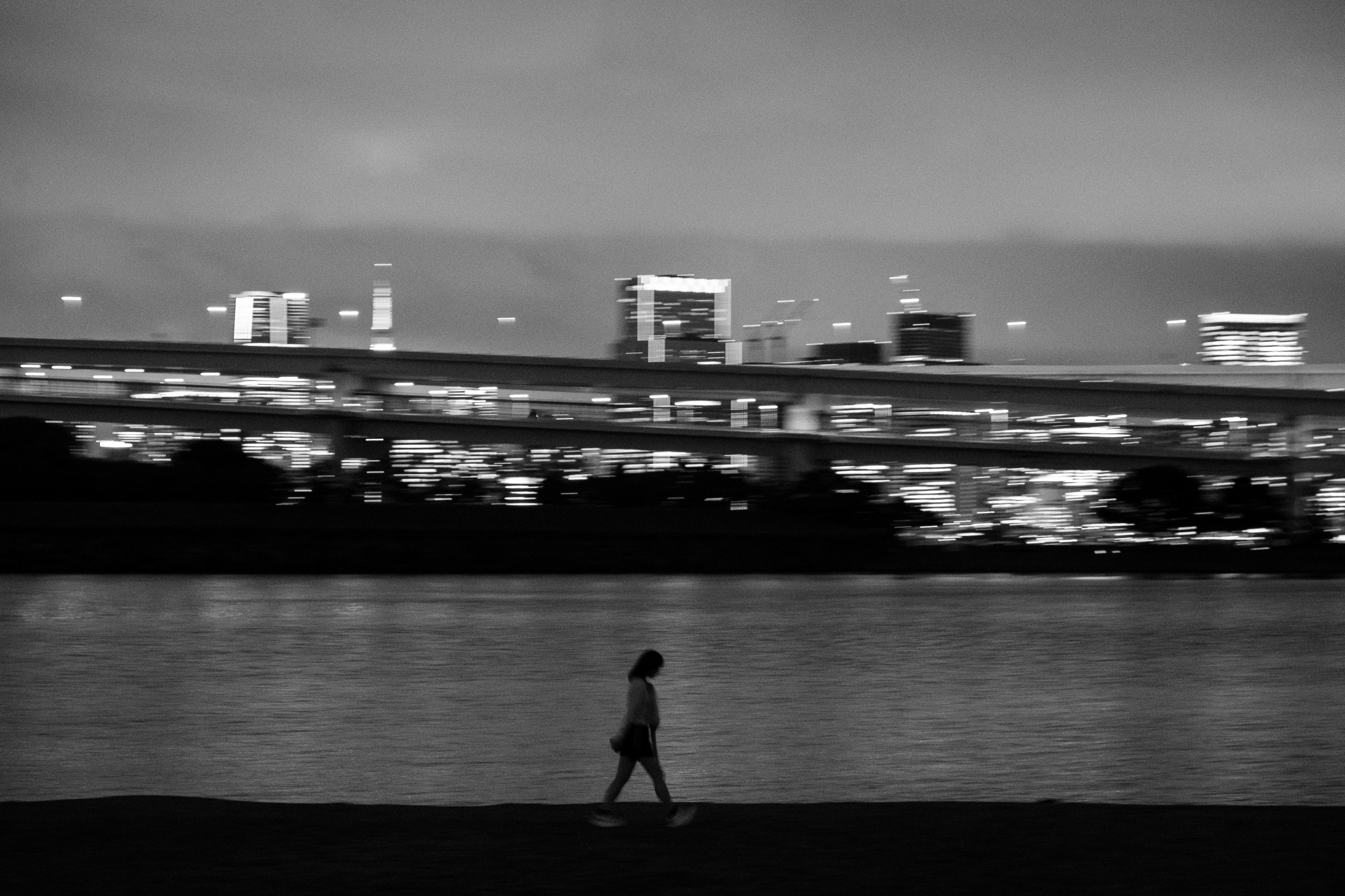 A lone figure walks along a waterfront at night, city lights blurred across the bay, black and white.