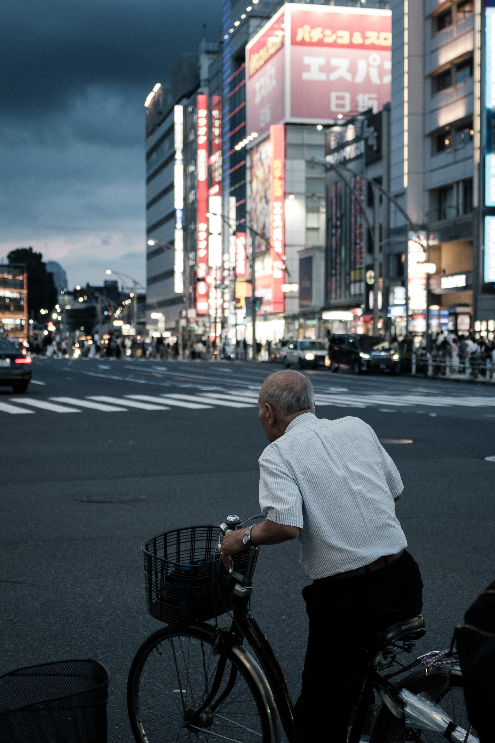 An elderly man on a bicycle, seen from behind, waits at a wide crossing lined with neon signs.