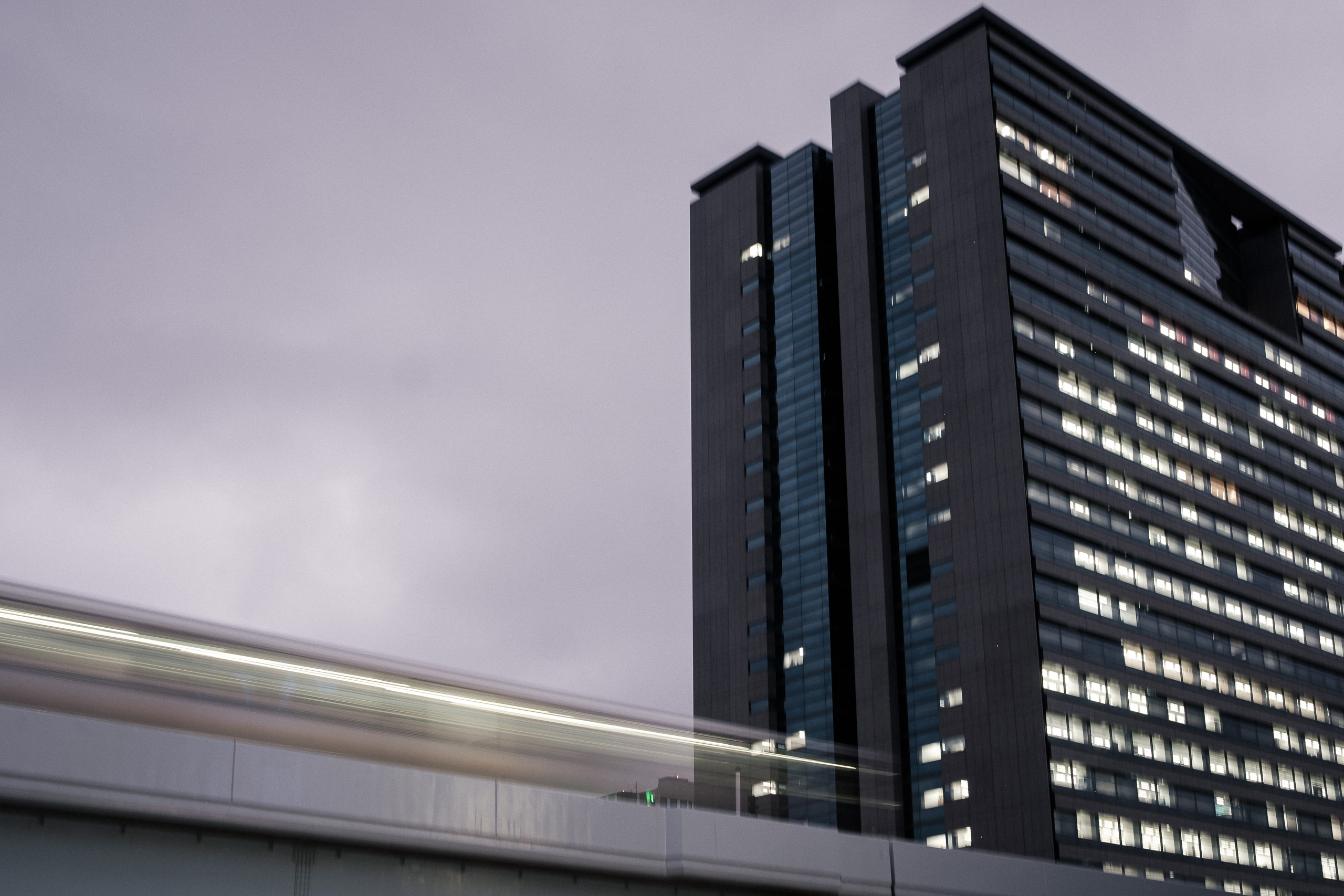 A light trail from a passing train sweeps past a lit office tower under a violet dusk sky.