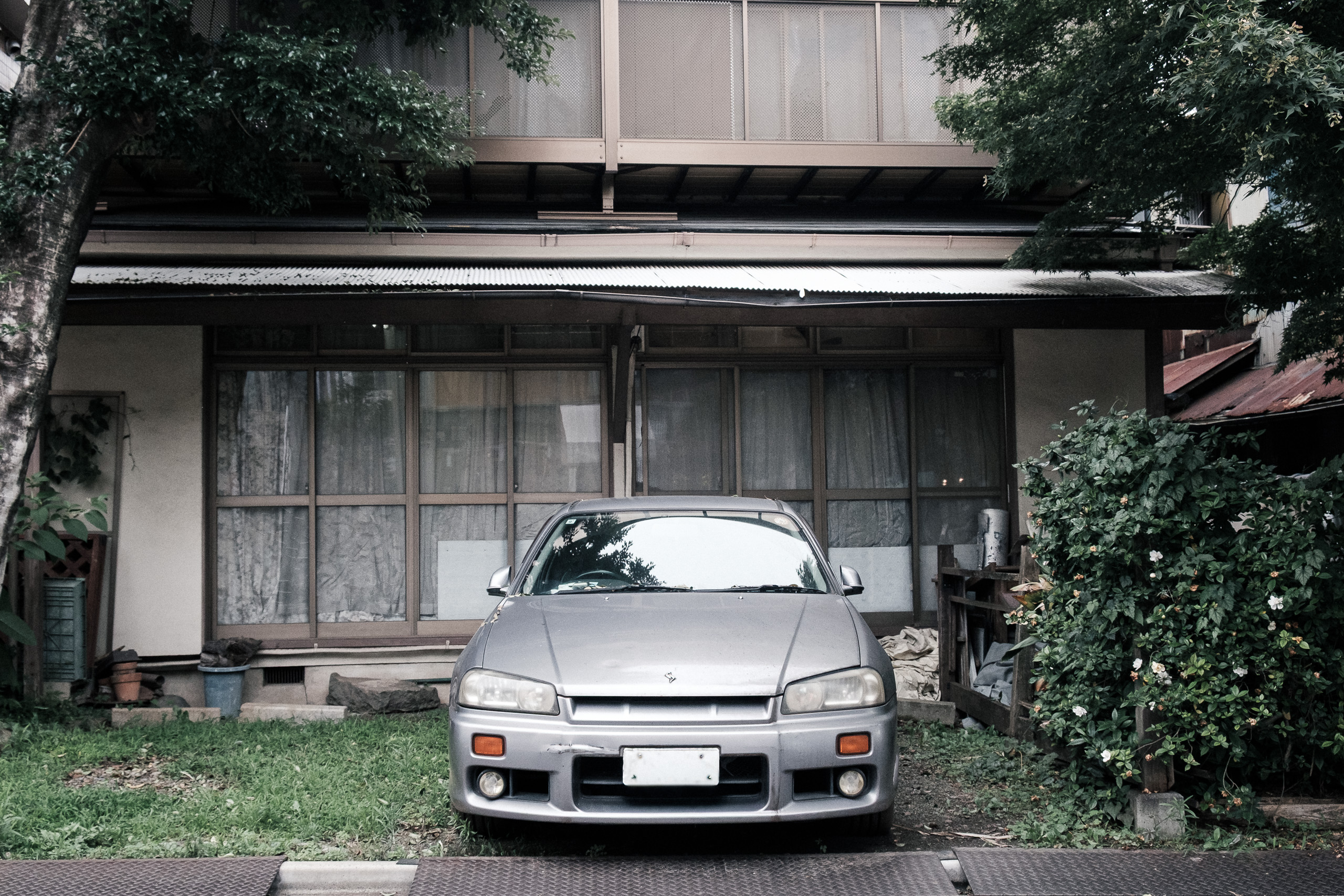 A silver car parked in front of an old wooden Japanese house, framed by trees.