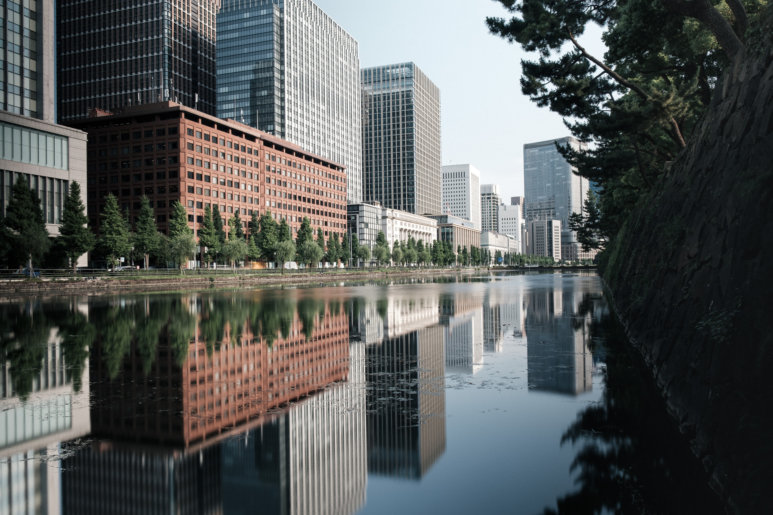 Office buildings reflected in the still water of a moat beside a dark stone wall.