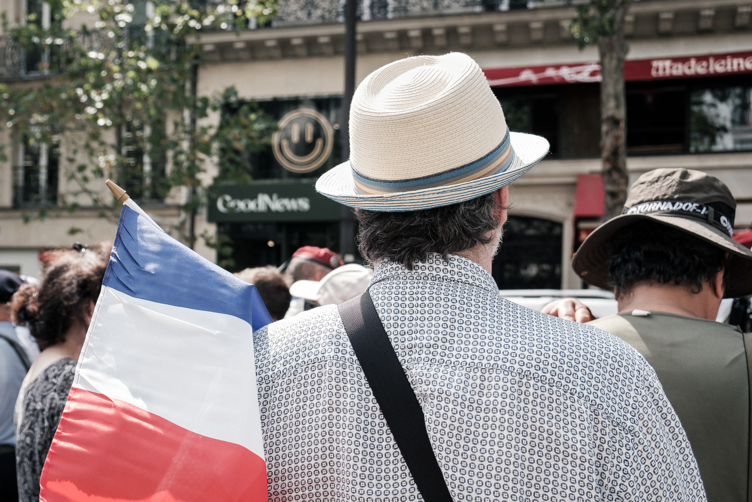 Man in a straw hat seen from behind in a crowd, a French tricolour flag beside him.