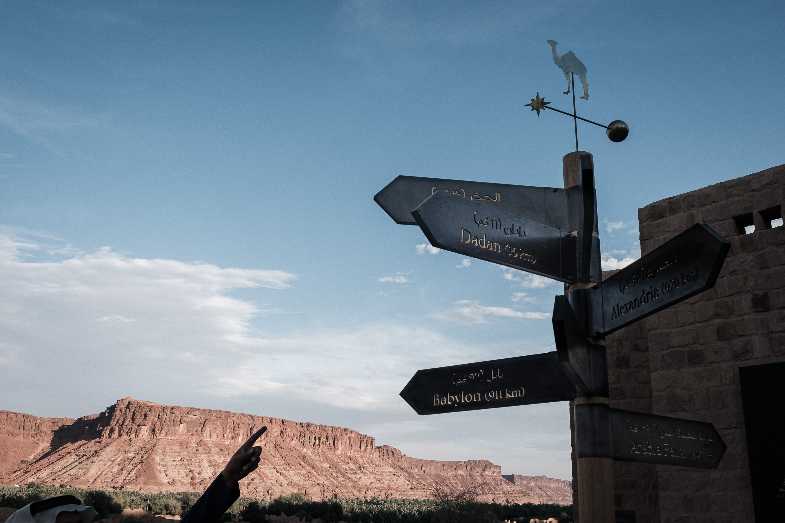 Wooden signpost with a camel weather vane and engraved signs pointing to Dadan, Alexandria and Babylon, with a hand pointing toward a sandstone cliff, AlUla, Saudi Arabia