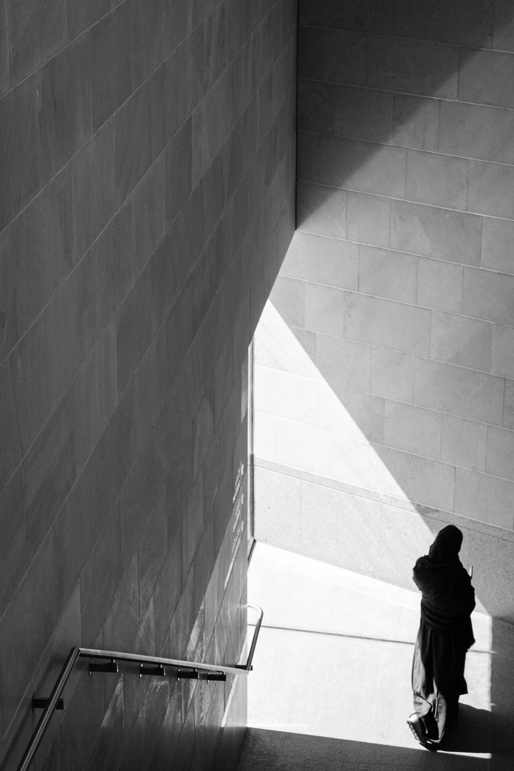 A figure in a long dark robe walking through a sharp diagonal of sunlight along a concrete wall, Riyadh, black and white