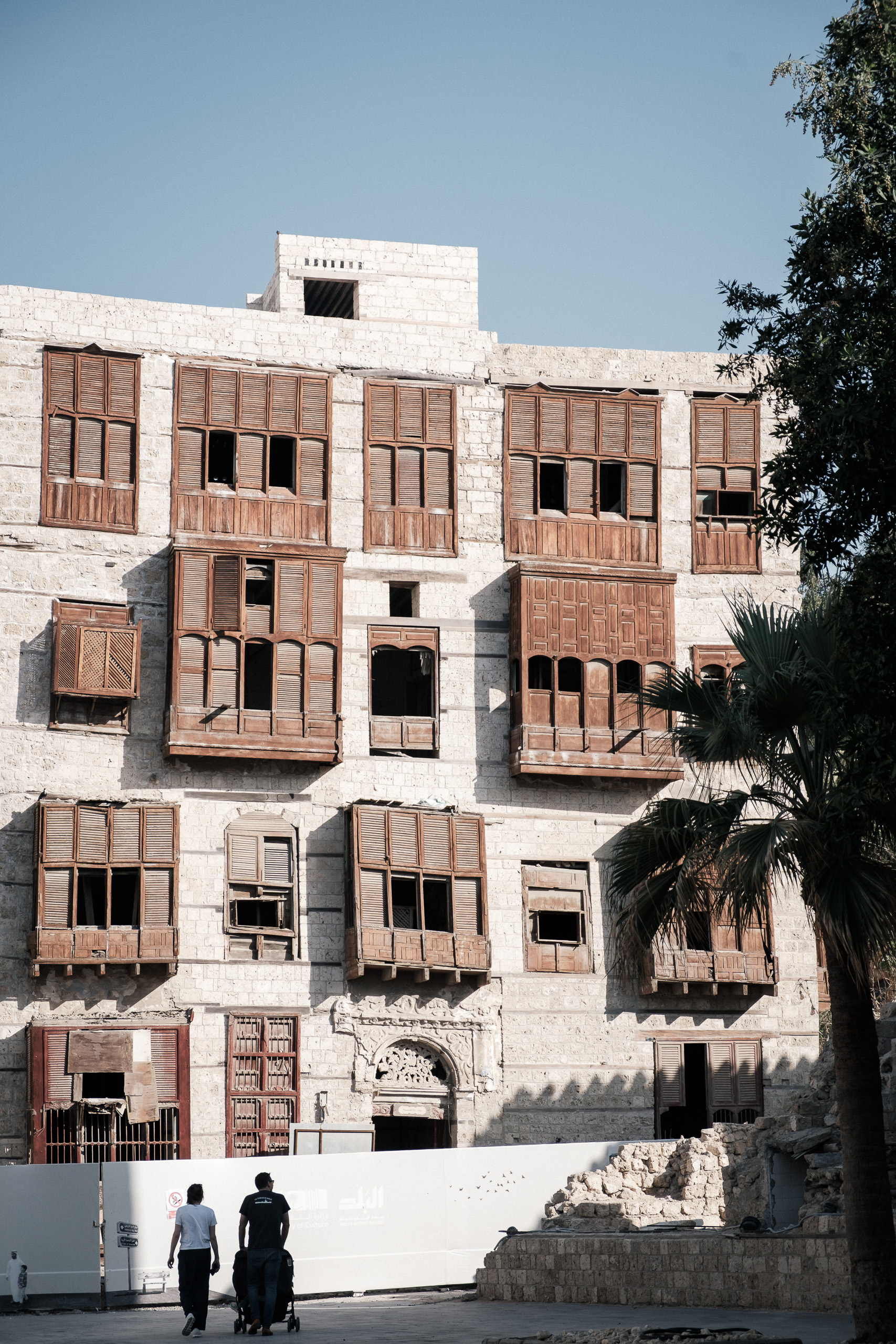 Weathered building with carved wooden windows in old Jeddah, two people walking away with a stroller