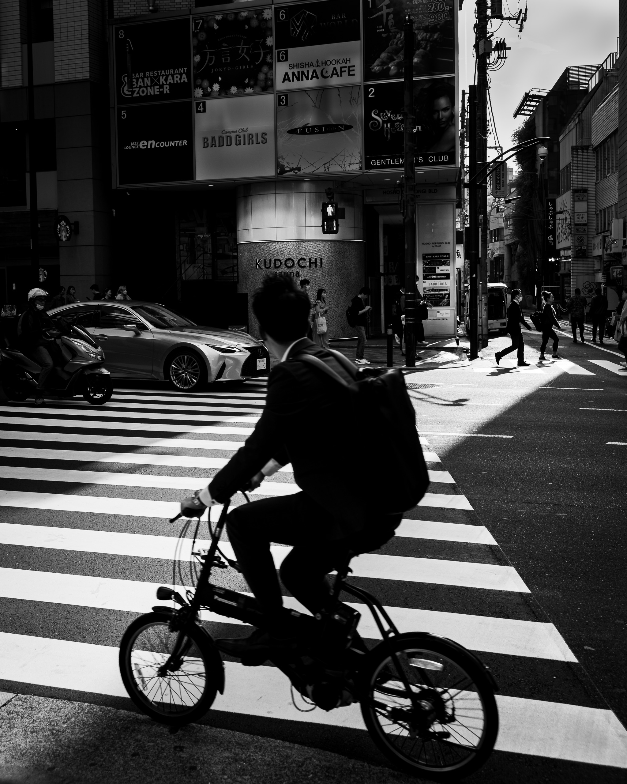A man in a suit with a backpack rides a small folding bicycle across a sunlit zebra crossing, with cars and pedestrians behind him.