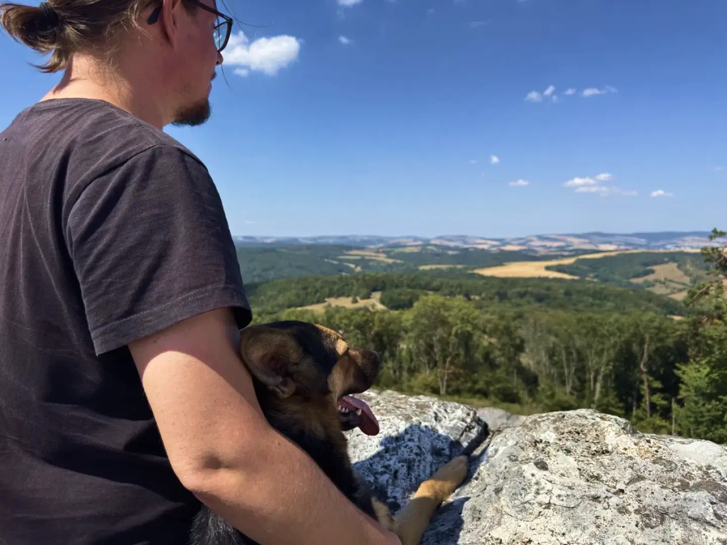 Mike sitting on a rocky lookout with his arm around Nova, a black and tan dog, overlooking forested hills