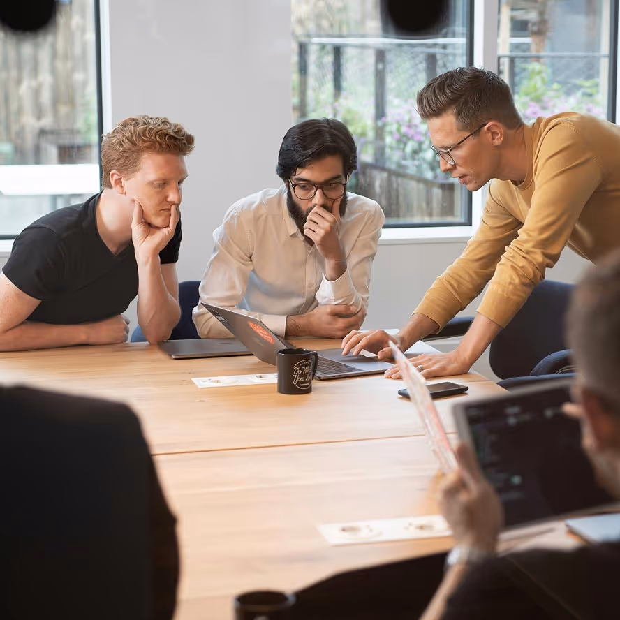 Three men in a meeting room focused on a laptop screen, with one standing and pointing at the laptop keyboard while two others sit and watch attentively.