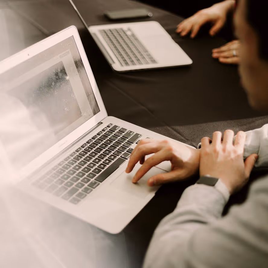 Person using a laptop on a dark table with another closed laptop and a smartphone nearby.