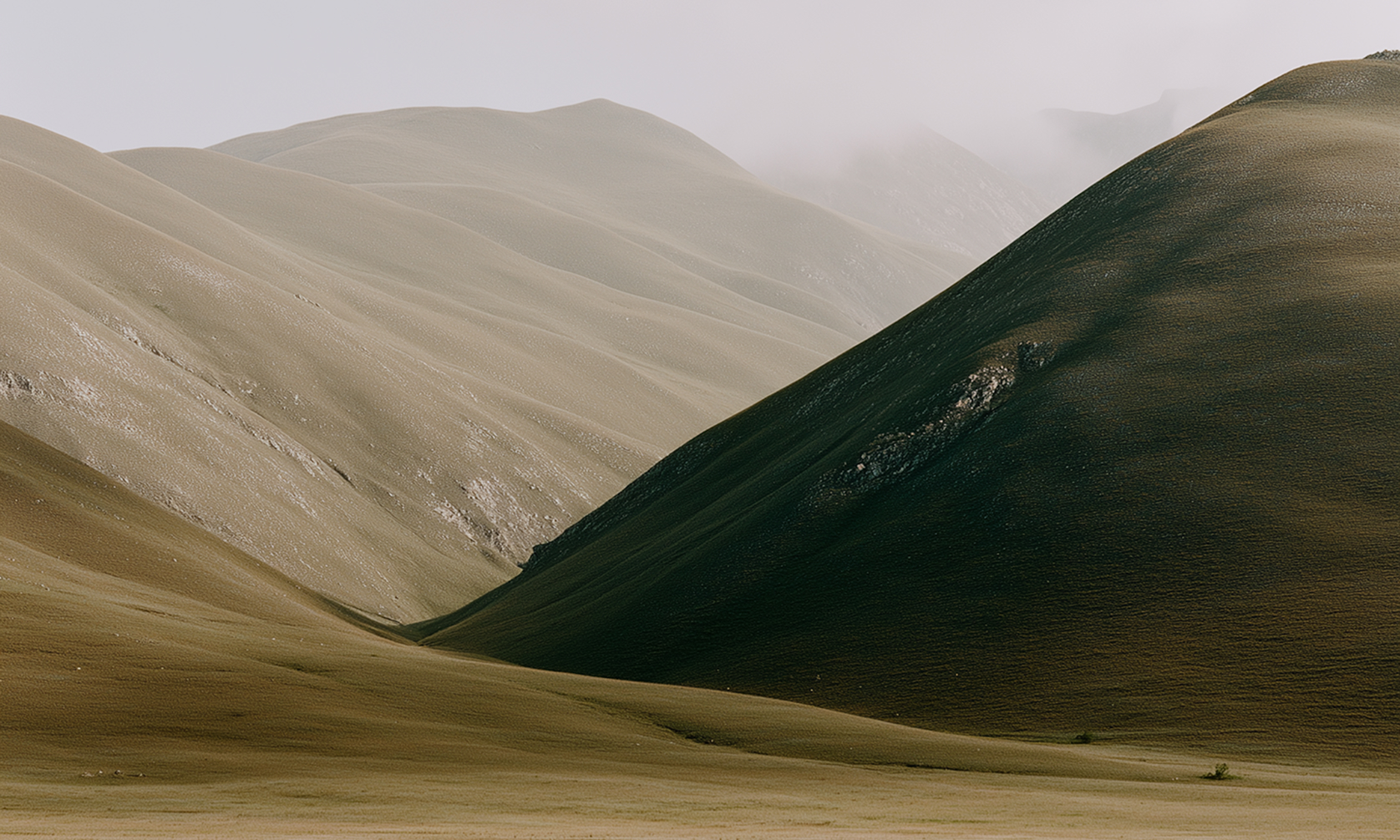 Gentle rolling hills with muted earthy tones under a foggy sky, showing a contrast between shaded and sunlit slopes and a wide valley in the foreground.