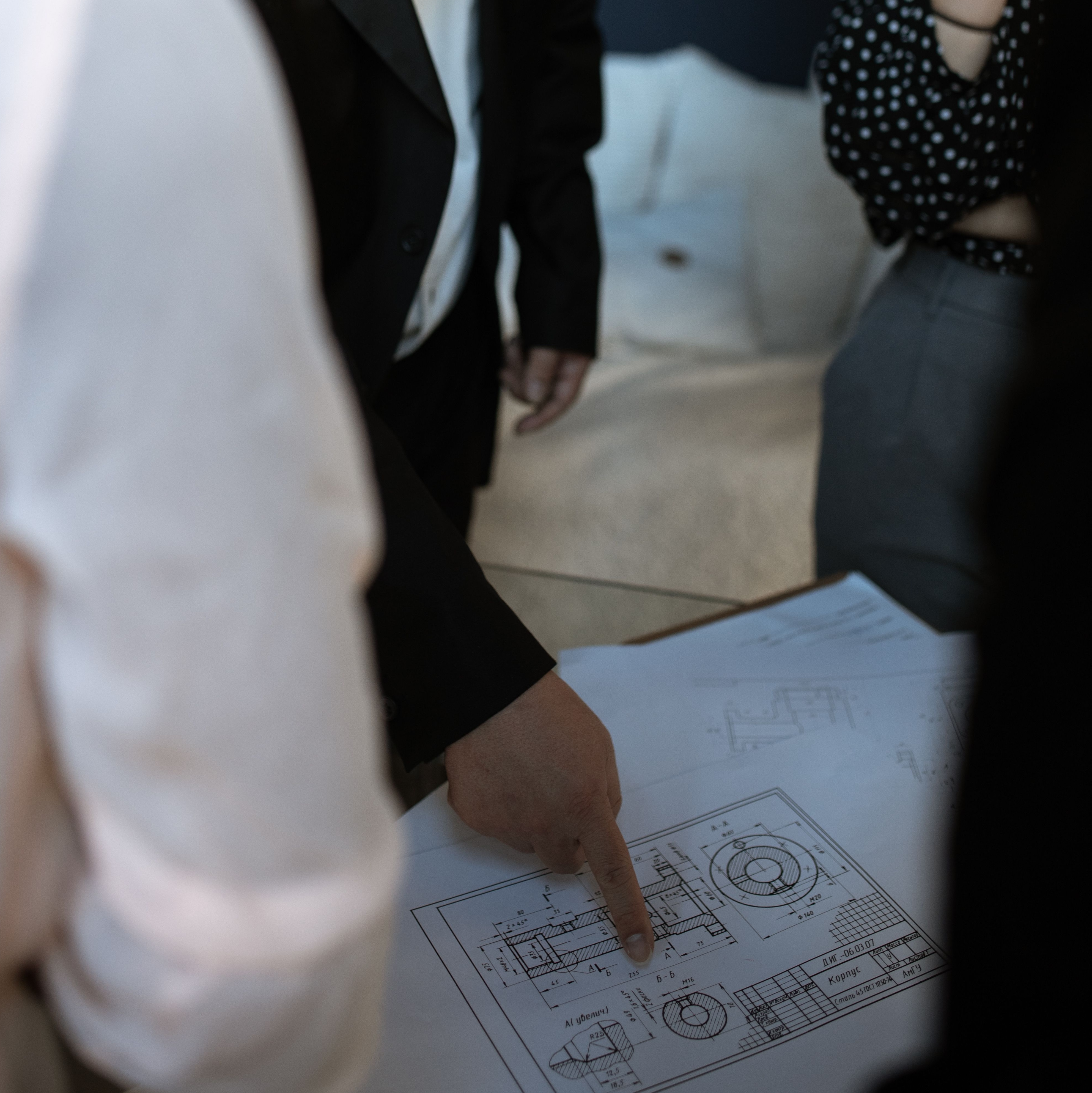 A person in a black suit points to a detailed technical drawing or blueprint on a table, while others stand nearby observing.