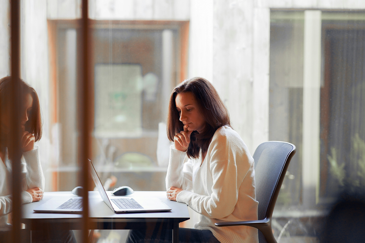 Woman in a white sweater sitting thoughtfully at a desk with a laptop in a bright office space, with her reflection visible on the glass beside her.