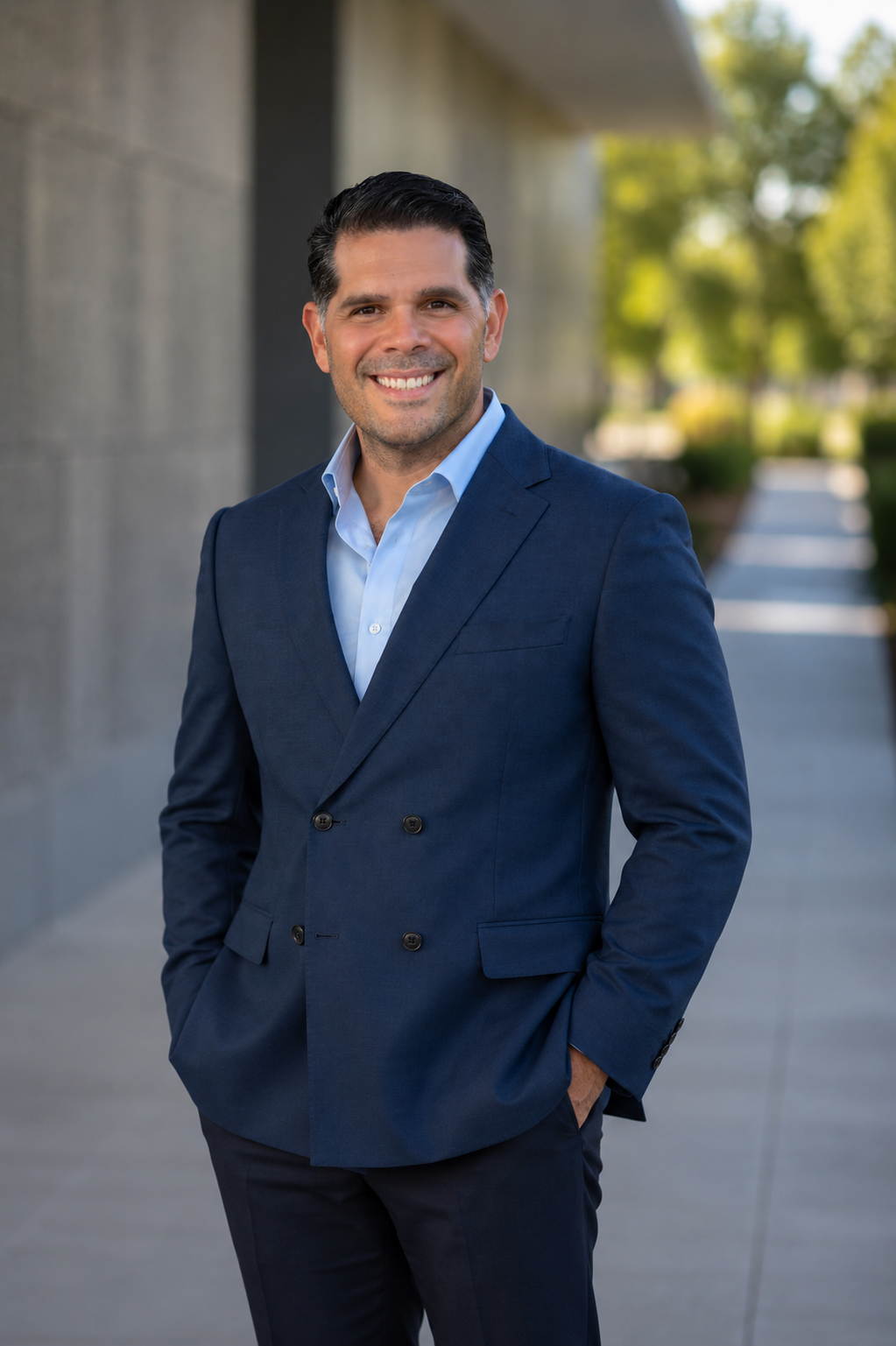 Smiling man with dark hair wearing a navy blue double-breasted suit jacket and light blue shirt stands outdoors with hands in pockets and a blurred background of greenery and a building.