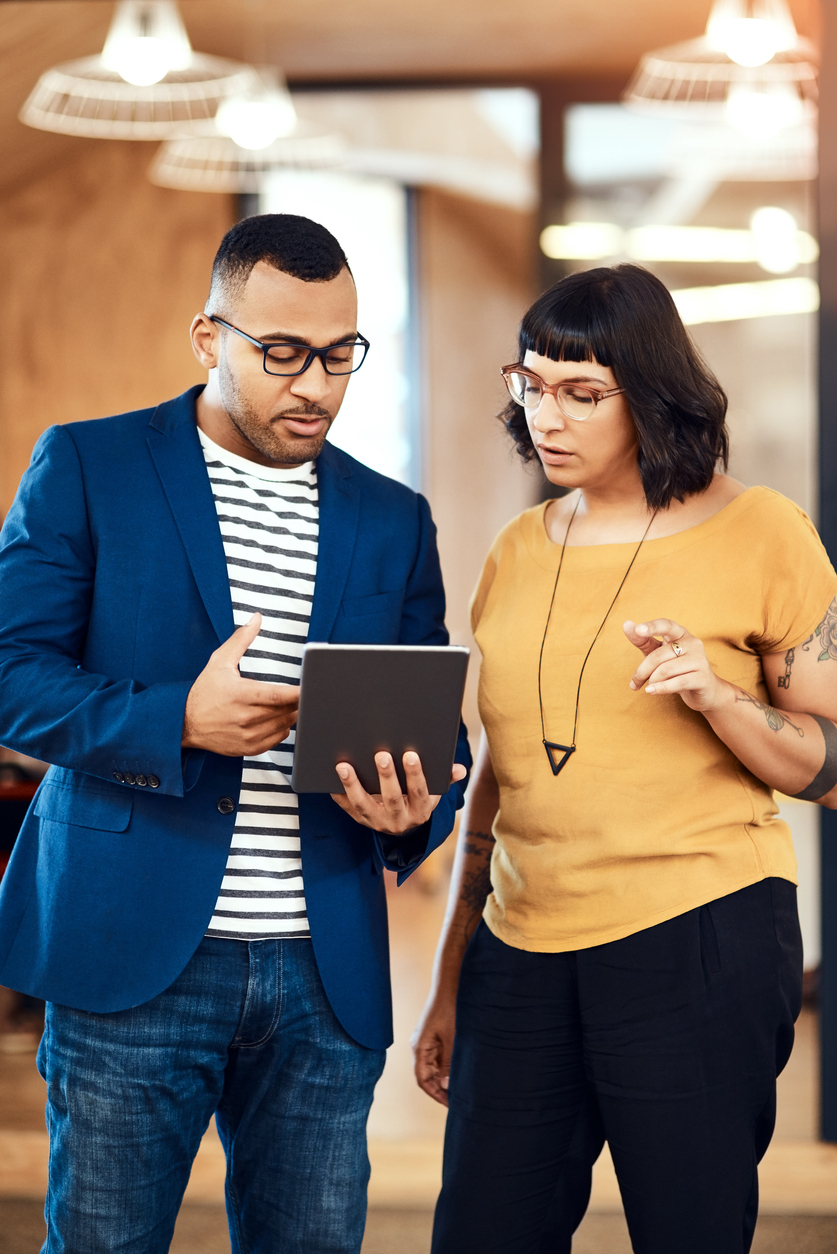 Two colleagues, a man in a blue blazer and striped shirt and a woman in a mustard yellow top, both wearing glasses, stand indoors engaged in discussion while looking at a tablet held by the man.