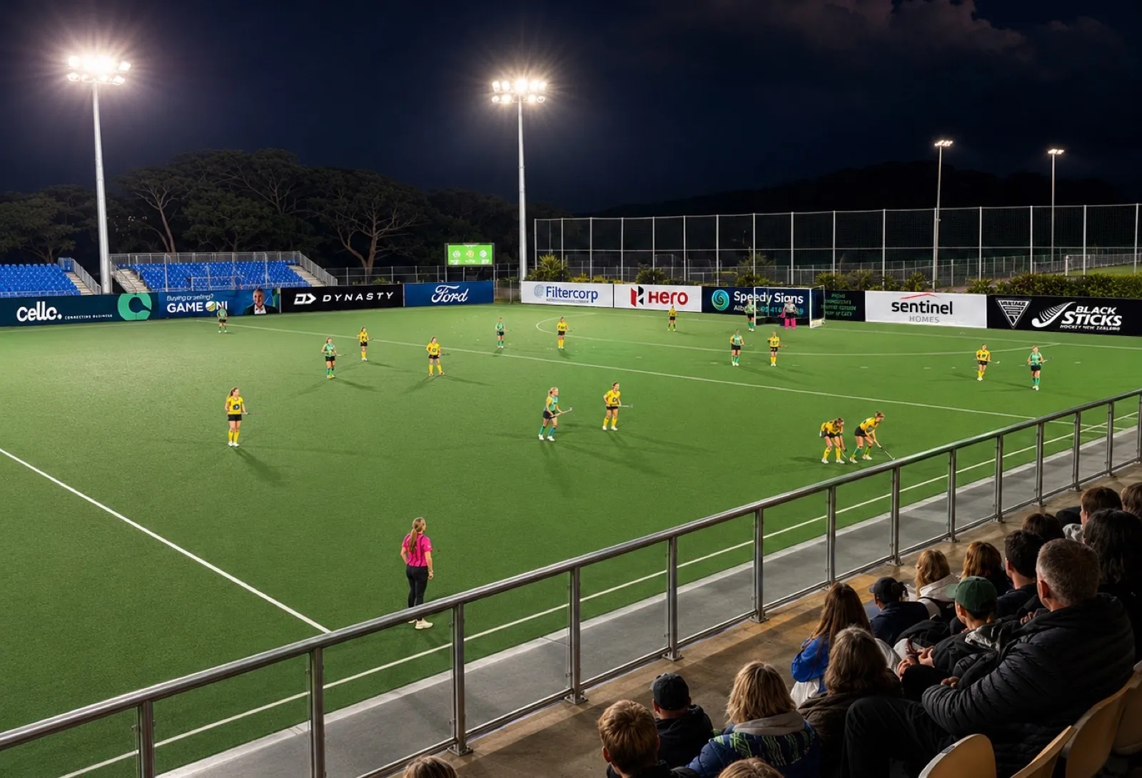 A night match at North Harbour Hockey, sponsor boards around the turf and spectators watching from the stand