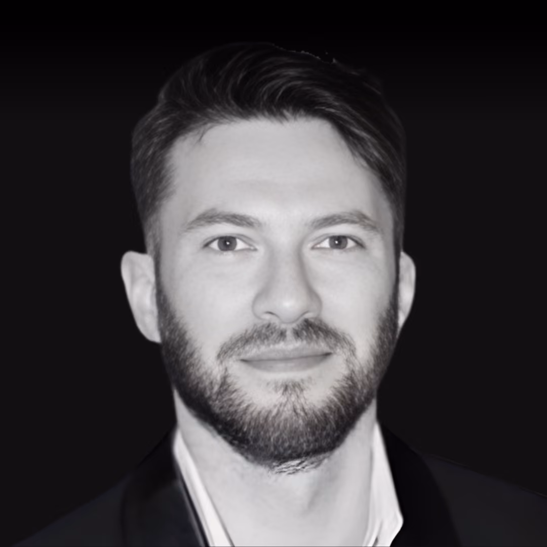 Portrait of a man with dark hair and a beard, wearing a dark jacket and light-colored shirt, against a black background.