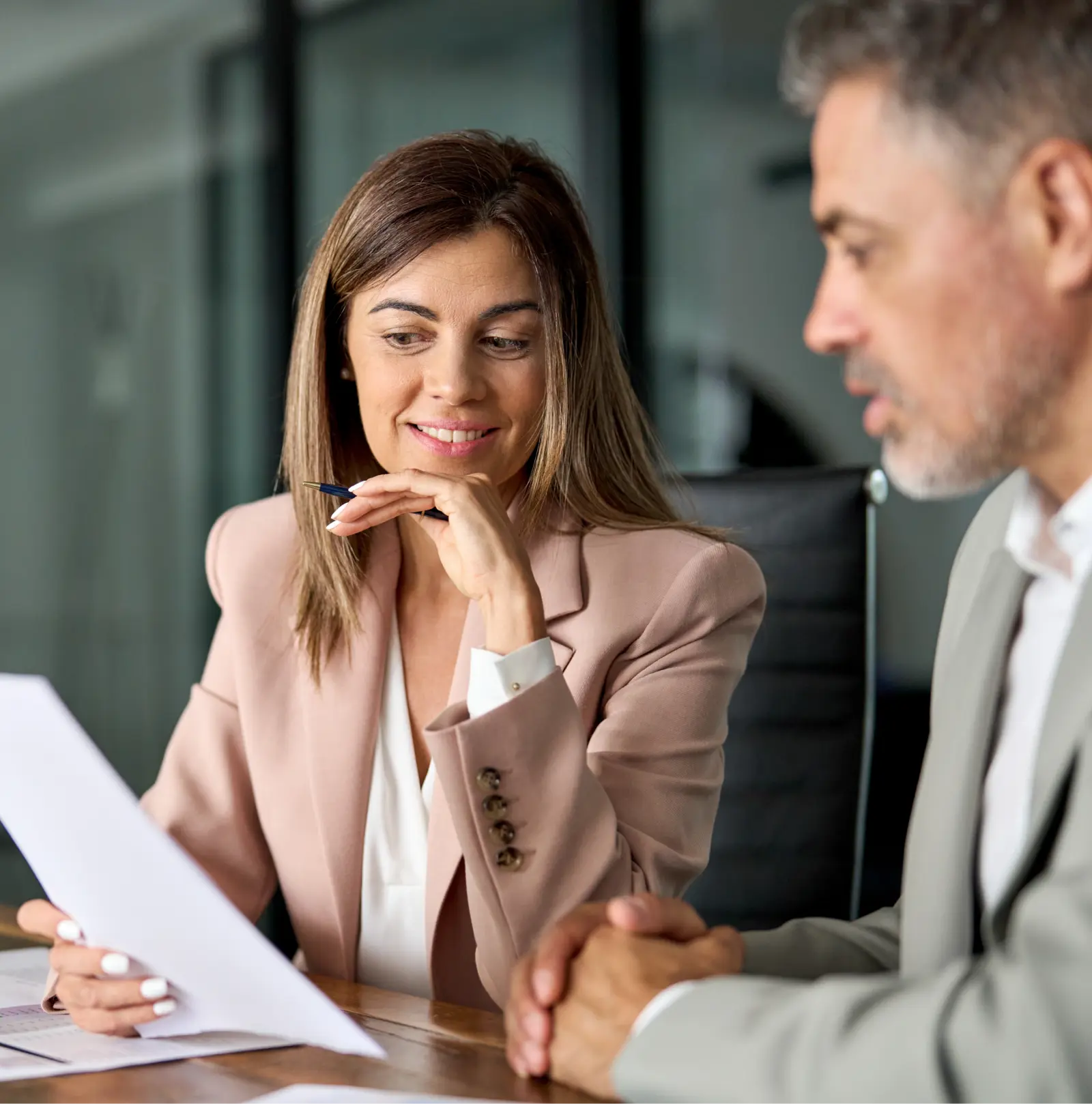 Woman in beige blazer reviewing a document while a man in grey suit listens attentively.