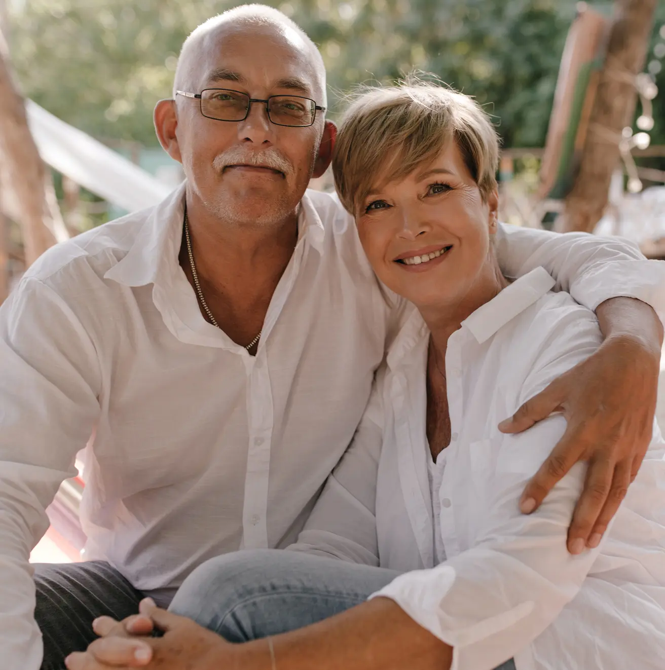 Smiling middle-aged couple in white shirts sitting outdoors with the man hugging the woman.