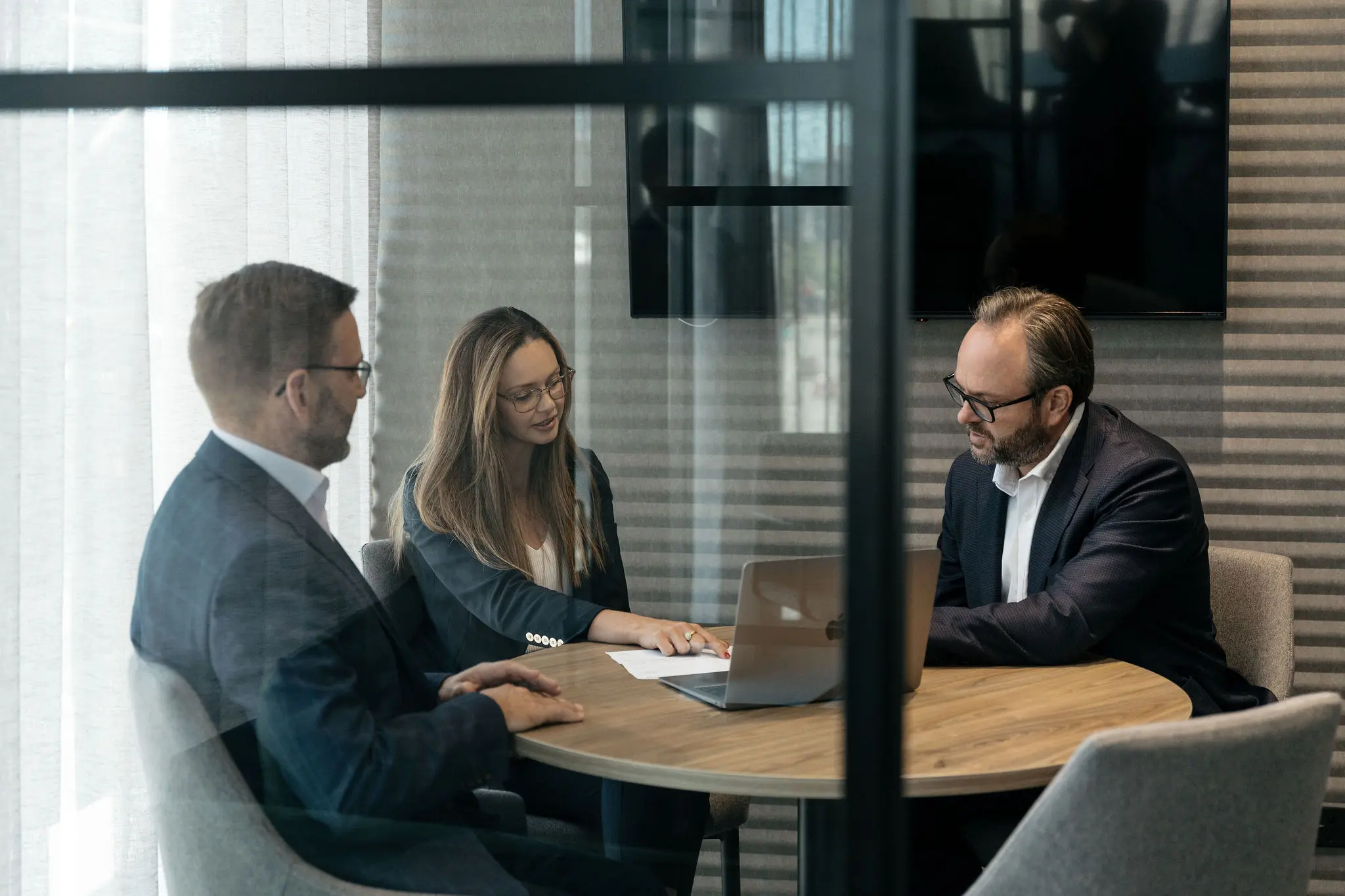 Three business professionals in a meeting room reviewing documents and a laptop at a round table.