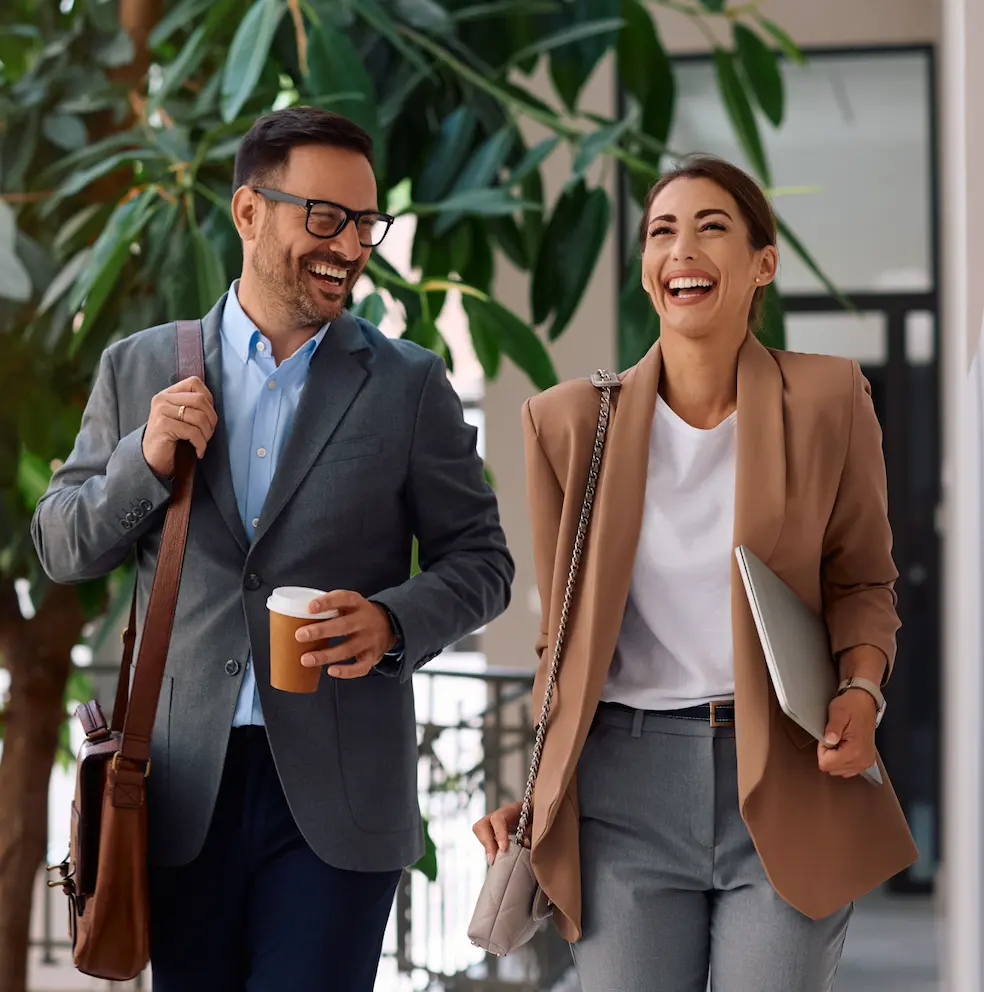 Smiling businessman and businesswoman walking outdoors, carrying a coffee and a laptop.