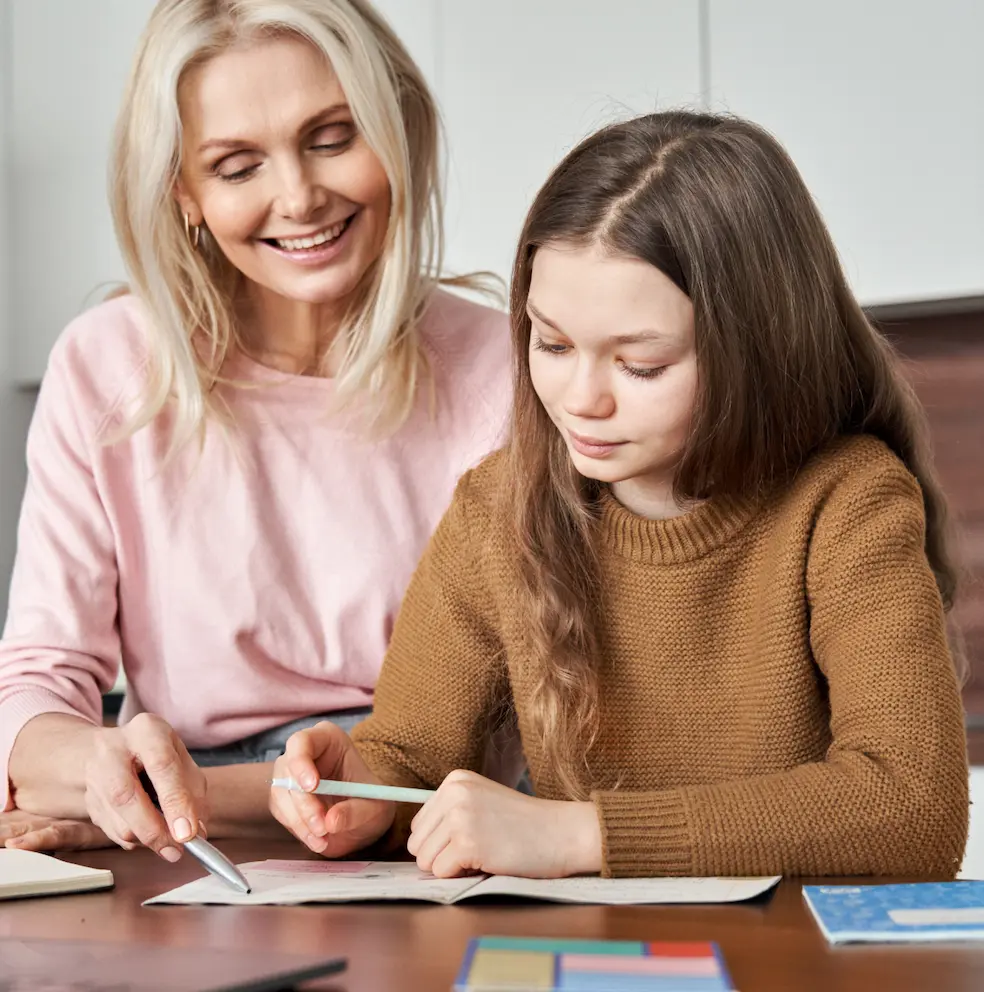Smiling woman helping a young girl with schoolwork at a table.