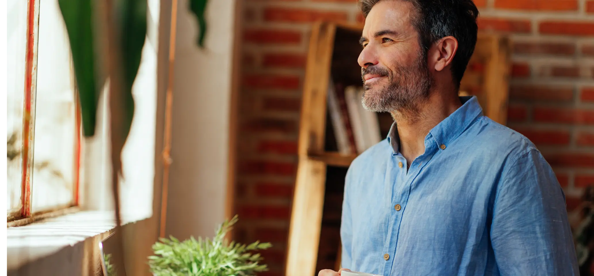 Smiling man in a blue shirt looking out a window while holding a coffee cup.